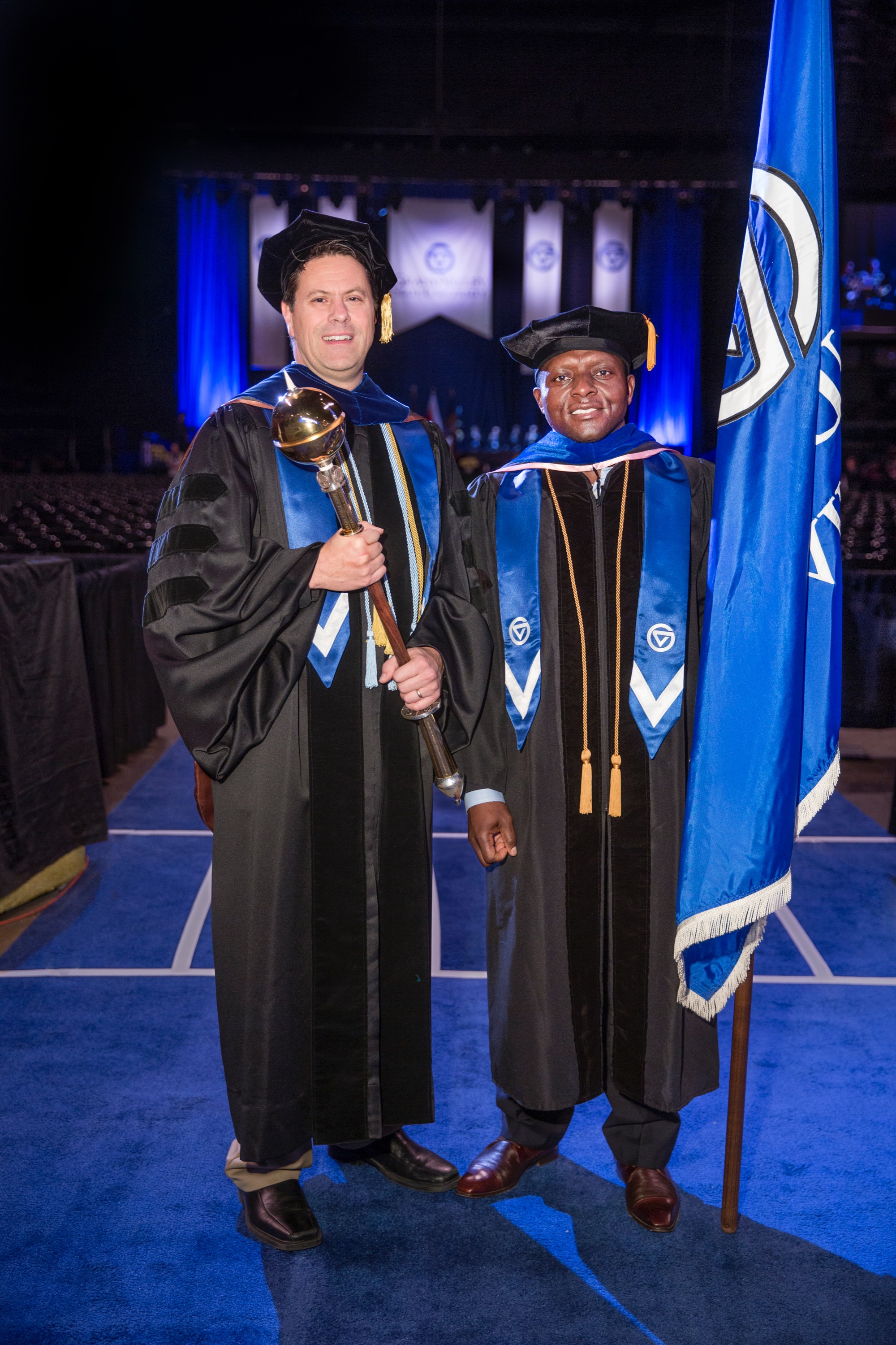 Two faculty members smiling. One holding the GVSU Mace and the other holding a GVSU flag